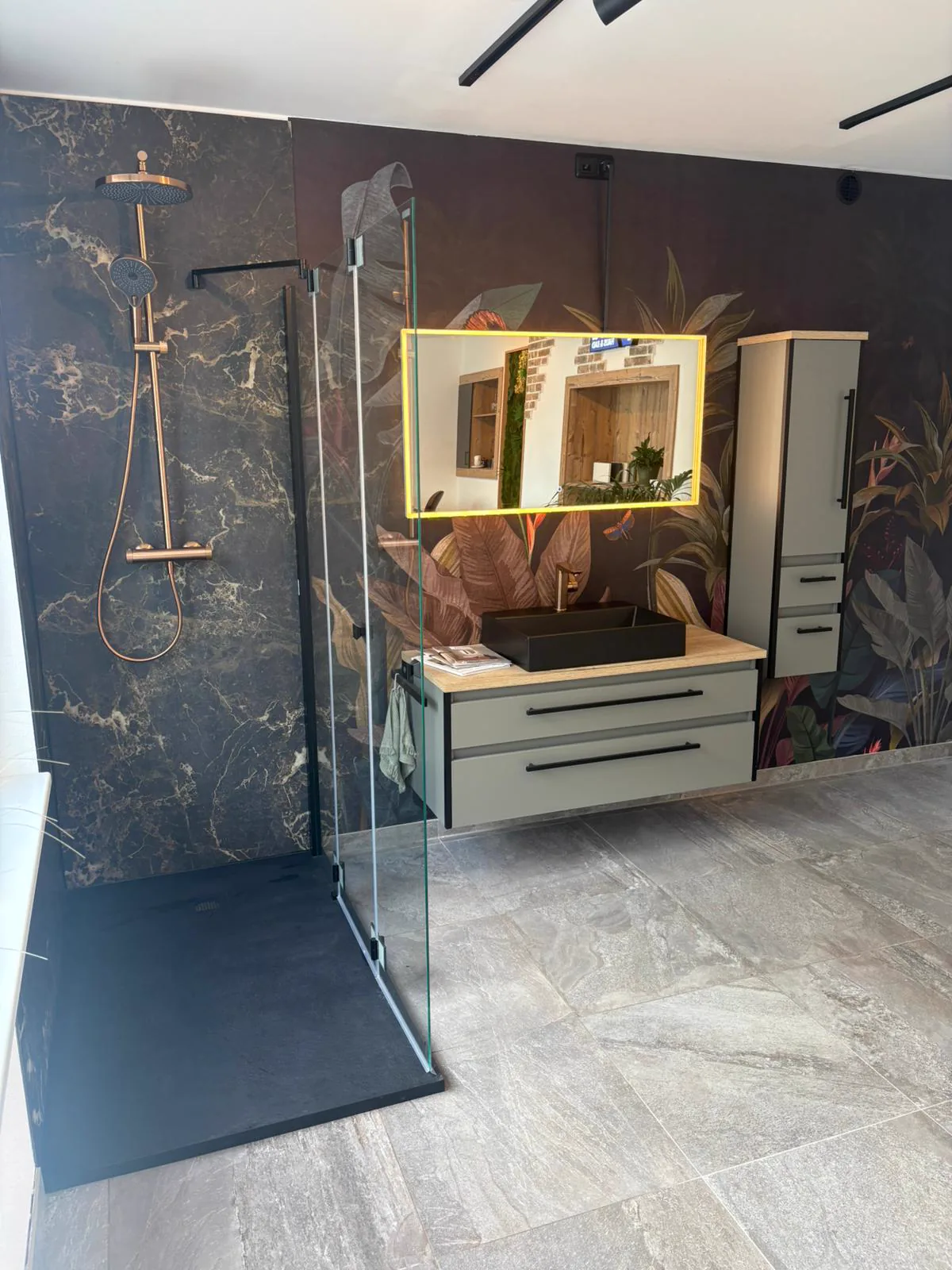 Ebenerdige Dusche mit dunklen Steinoptik-Paneelen und goldener Regenbrause im Schauraum von FoFa Haus & Bad